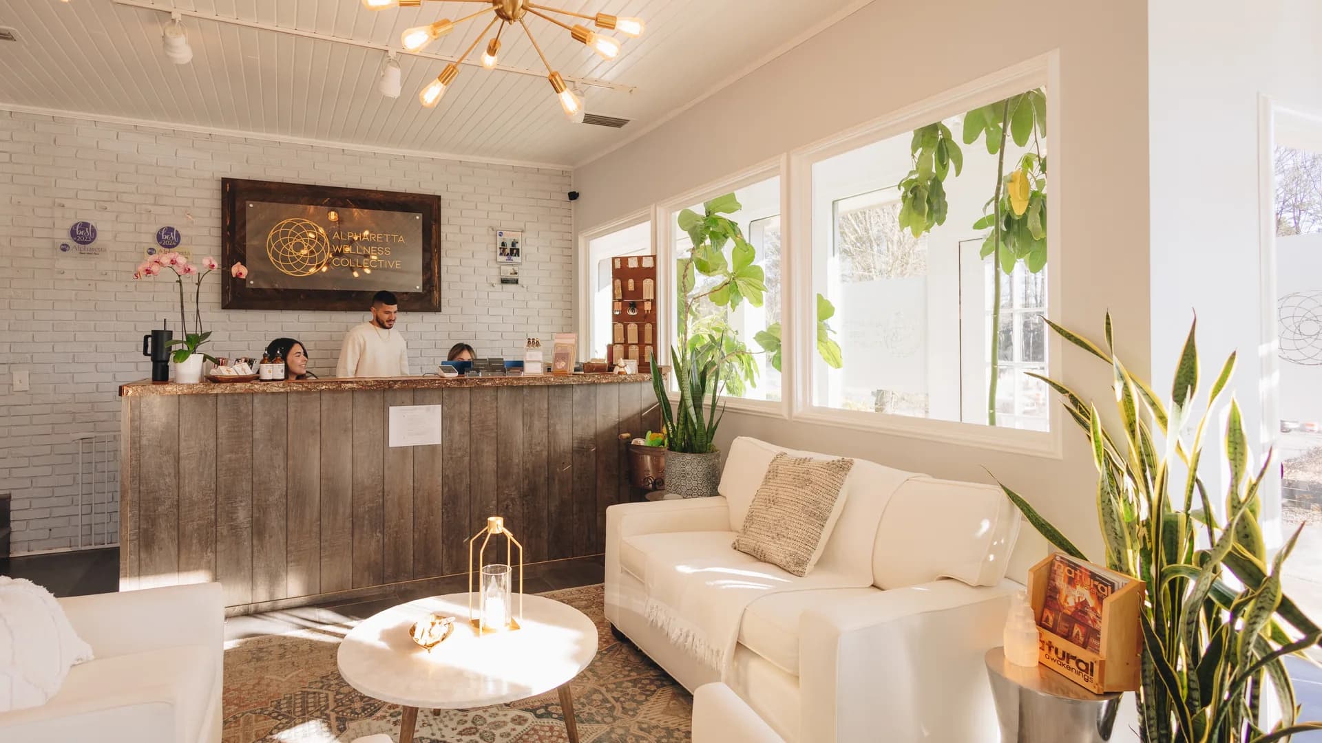 The reception room at Alpharetta Wellness Collective: white armchairs and a low table in front of the reclaimed-wood desk, with two of the team behind it.