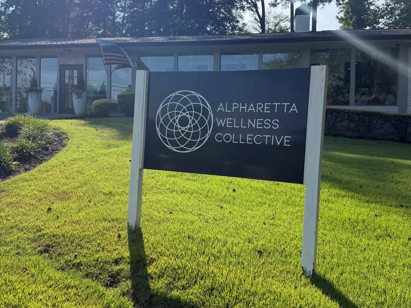 The dark Alpharetta Wellness Collective sign on white posts on the lawn at North Main Street, with the front door of 264 behind it.