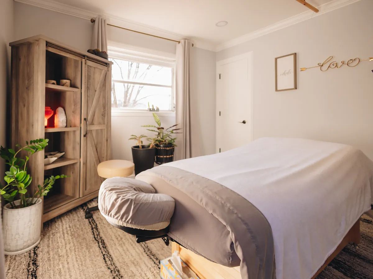 A treatment room made up for the next patient: a massage table under a white blanket, plants, and a salt lamp glowing in a wooden cabinet by the window.