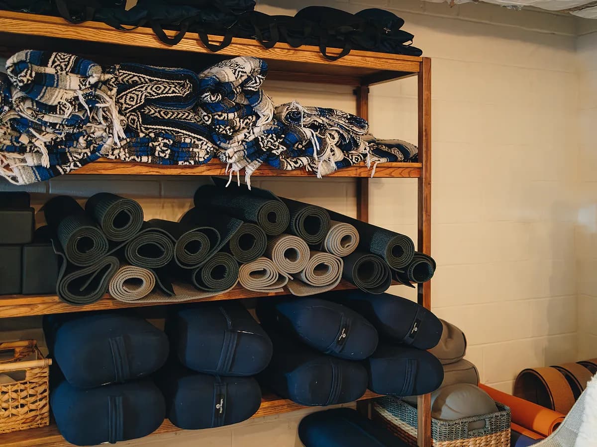A wooden shelf stacked with woven blankets, rolled yoga mats, blocks and navy bolsters.